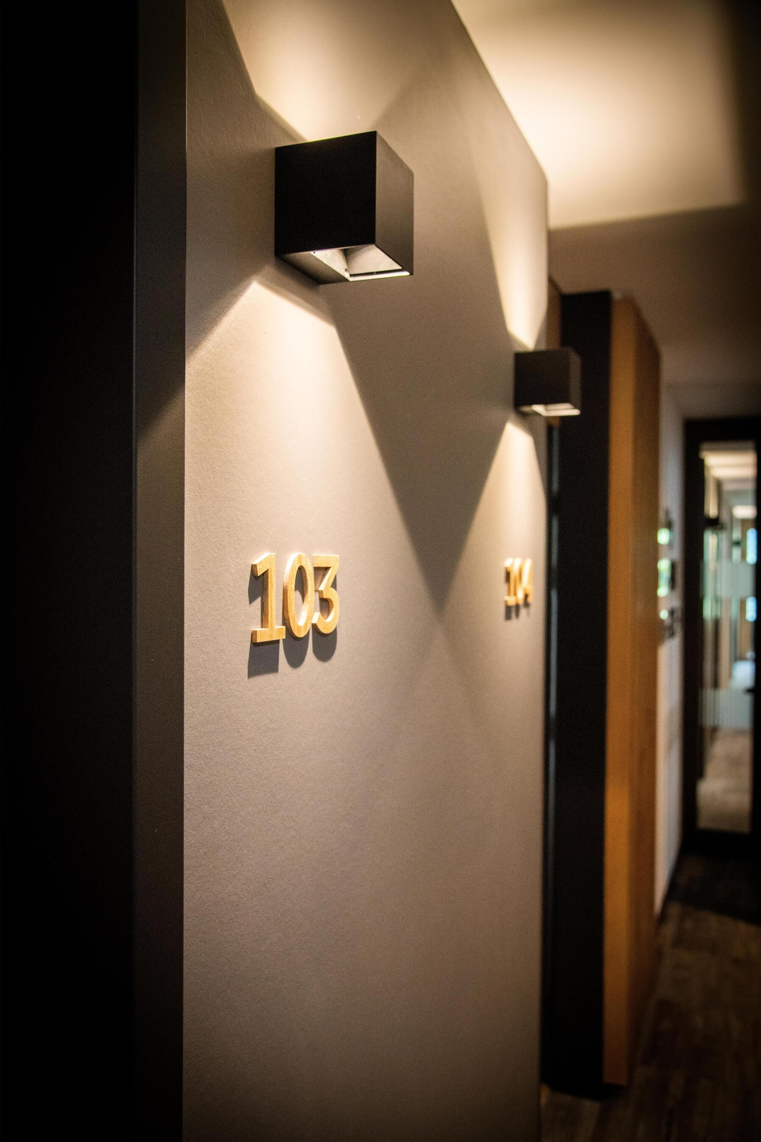 A hallway with illuminated room numbers '103' and '104' on the wall, a modern light fixture, and a wooden floor.