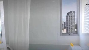 A modern bathroom with a bathtub, a window with a cityscape view, and a shower area. - My Resort BKK (Bangkok)