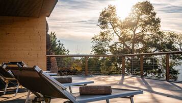 A rooftop terrace with sun loungers, a table, and a view of trees and a body of water.