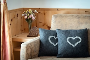 Ein Blick in einen Raum mit einer hölzernen Bank, einer Vase mit Blumen und einem Sofa mit herzmusterten Kissen.