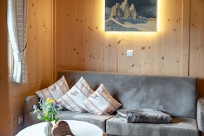 Ein Zimmer mit Holzwänden, einem grauen Sofa, gemusterten Kissen, einem gerahmten Landschaftsbild und einer Vase mit Blumen.