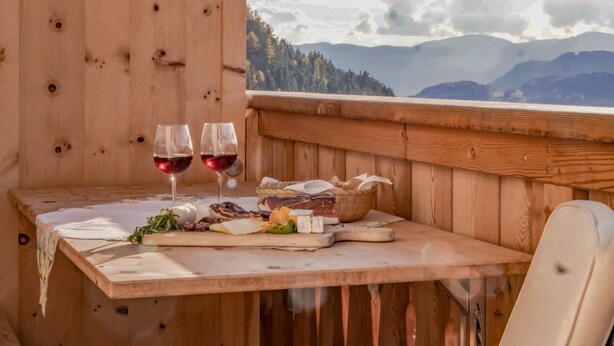 A wooden table with two glasses of red wine, a basket of bread, and a plate of cheese and vegetables.