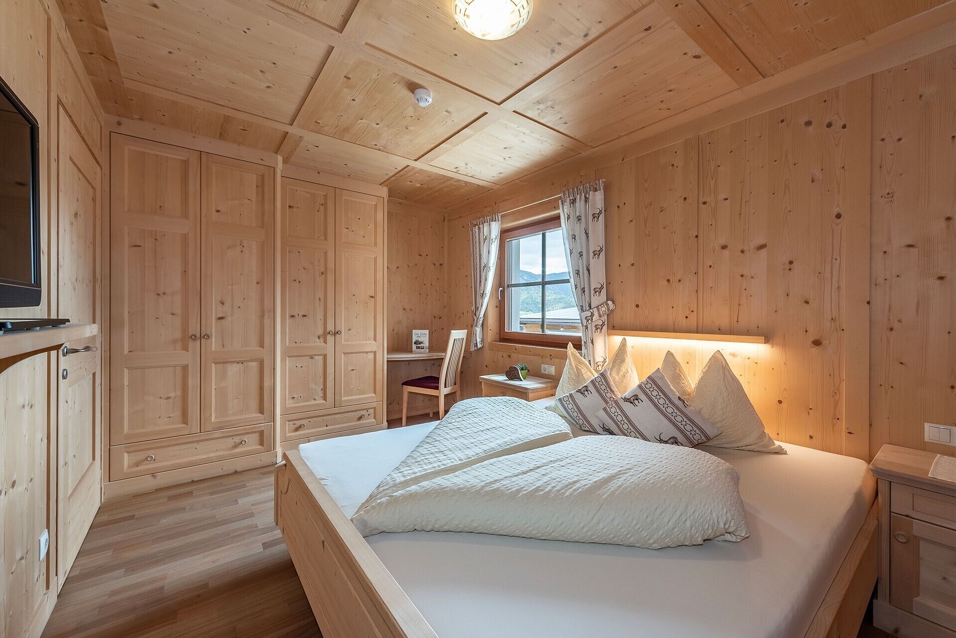 A wooden cabin bedroom with a bed, a desk, and a window with curtains.