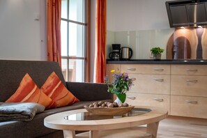 Un salon moderne avec un canapé, une table basse sur laquelle se trouve un bol de noix, et un coin cuisine équipé de placards et d’une machine à café.