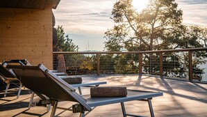 Une terrasse sur le toit, équipée de chaises longues, d’une table et offrant une vue sur les arbres et une étendue d’eau.