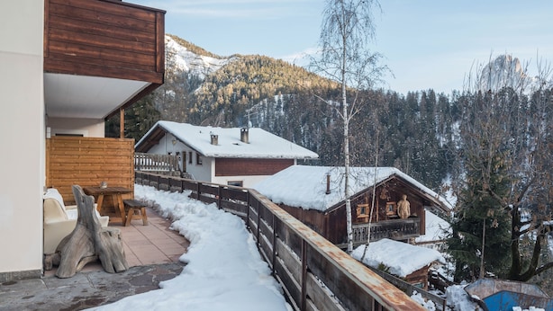 A wooden cabin with a snow-covered roof, a wooden deck, and a view of a forested mountain. - Cosy Apartment “Ulrica” with Mountain View, Wi-Fi, Balcony & Terrace (St. Ulrich / Gröden)