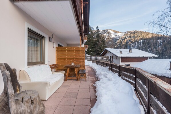 A wooden deck with a sofa and table, snow-covered ground, and a mountain view.
