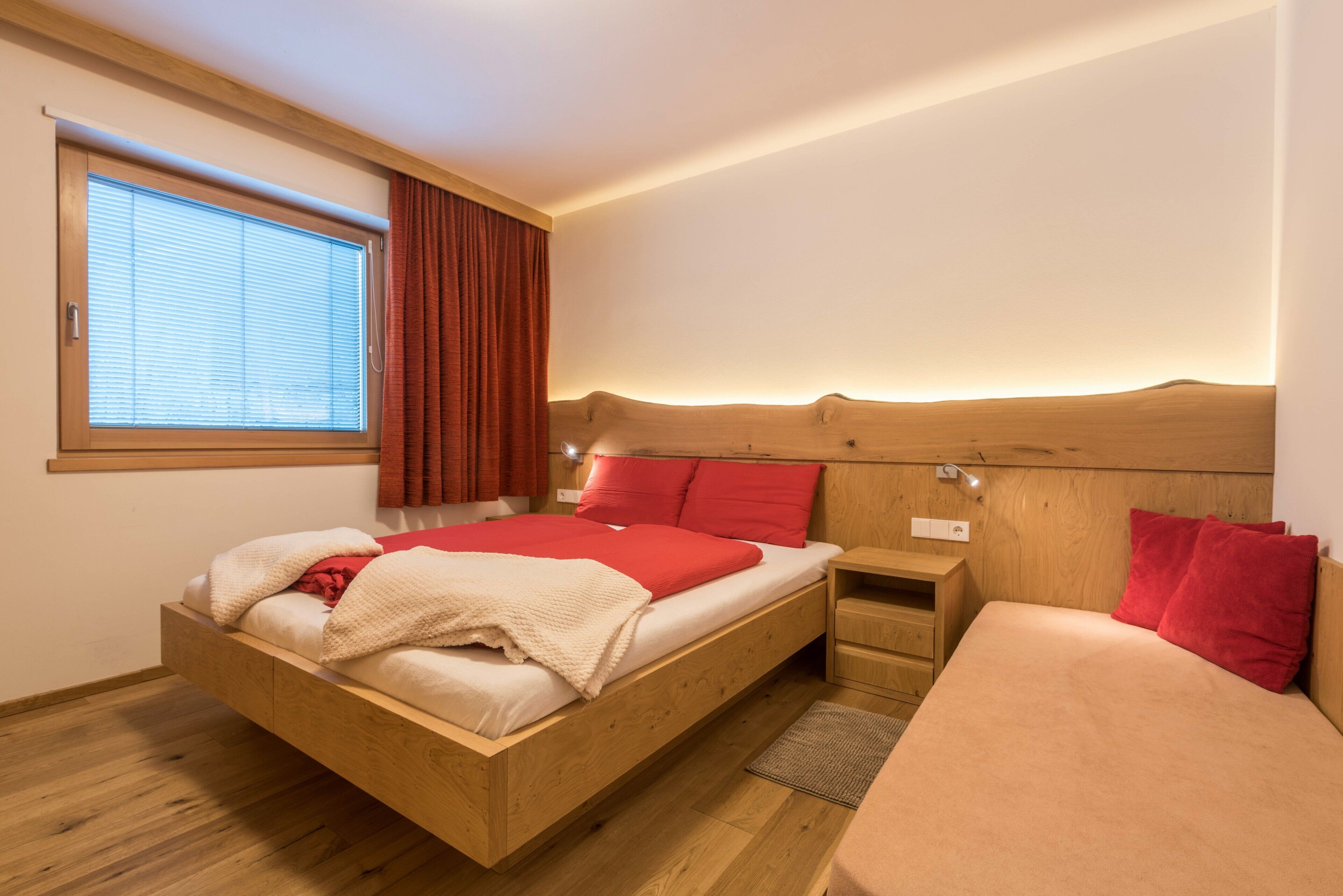 Ein hölzernes Bett mit roten Kissen, ein Nachttisch und ein Fenster mit Vorhängen.
