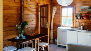 A wooden cabin interior with a dining table, benches, a kitchen area with a sink, and a window with curtains.