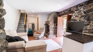 A living room with a stone wall, a sofa, a chair, a TV, and a fireplace. - House in the Mountains With Garden (São Vicente)