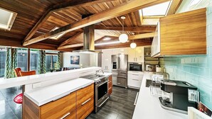 Une cuisine moderne avec des poutres en bois, un grand îlot central, des appareils électroménagers en acier inoxydable et une vue sur l’extérieur.