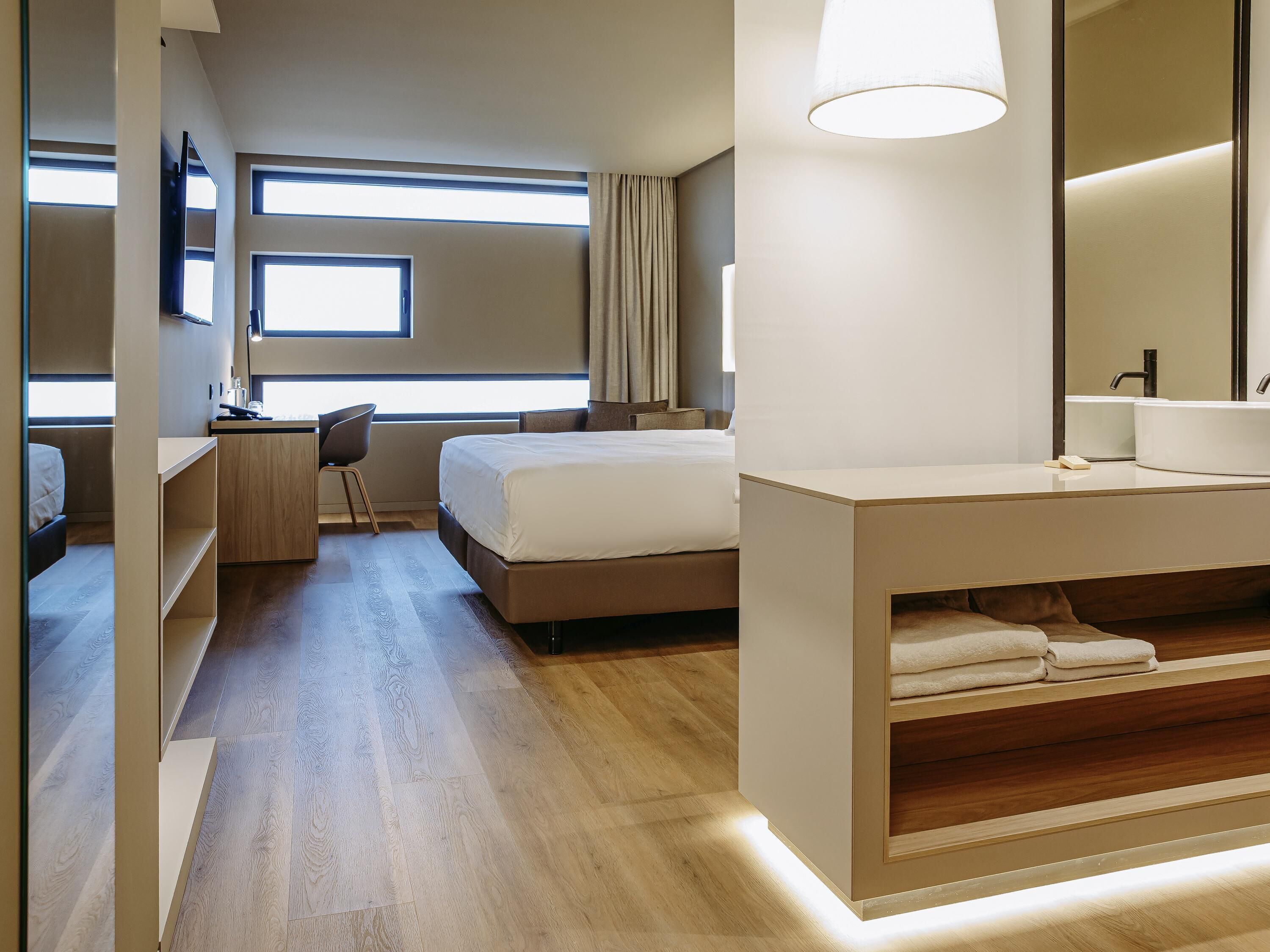 A modern hotel room with a bed, a desk, a chair, a large mirror, and a bathroom with a sink and towel storage.