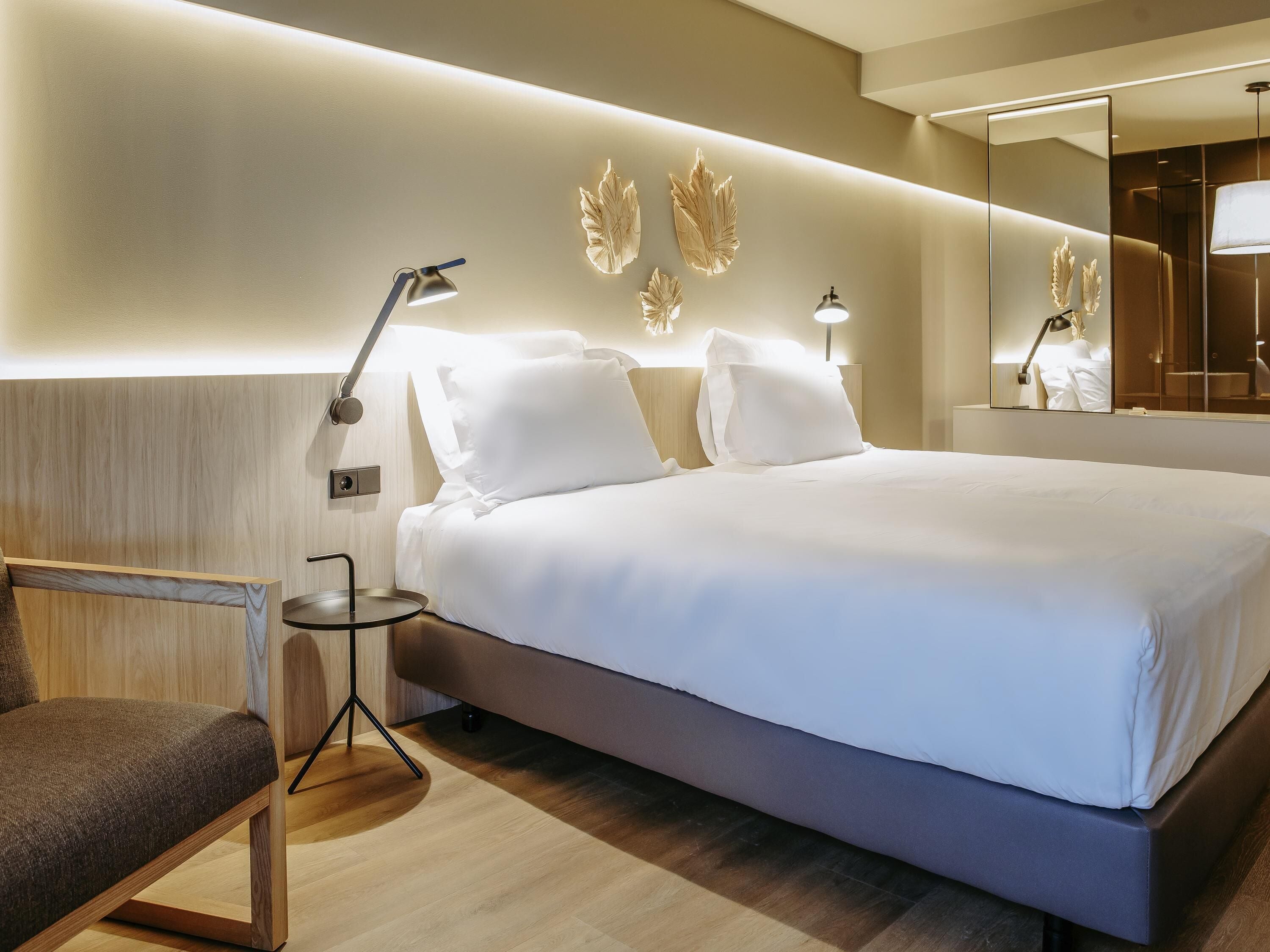 A modern hotel room with a large bed, bedside lamps, a chair, and a mirror.