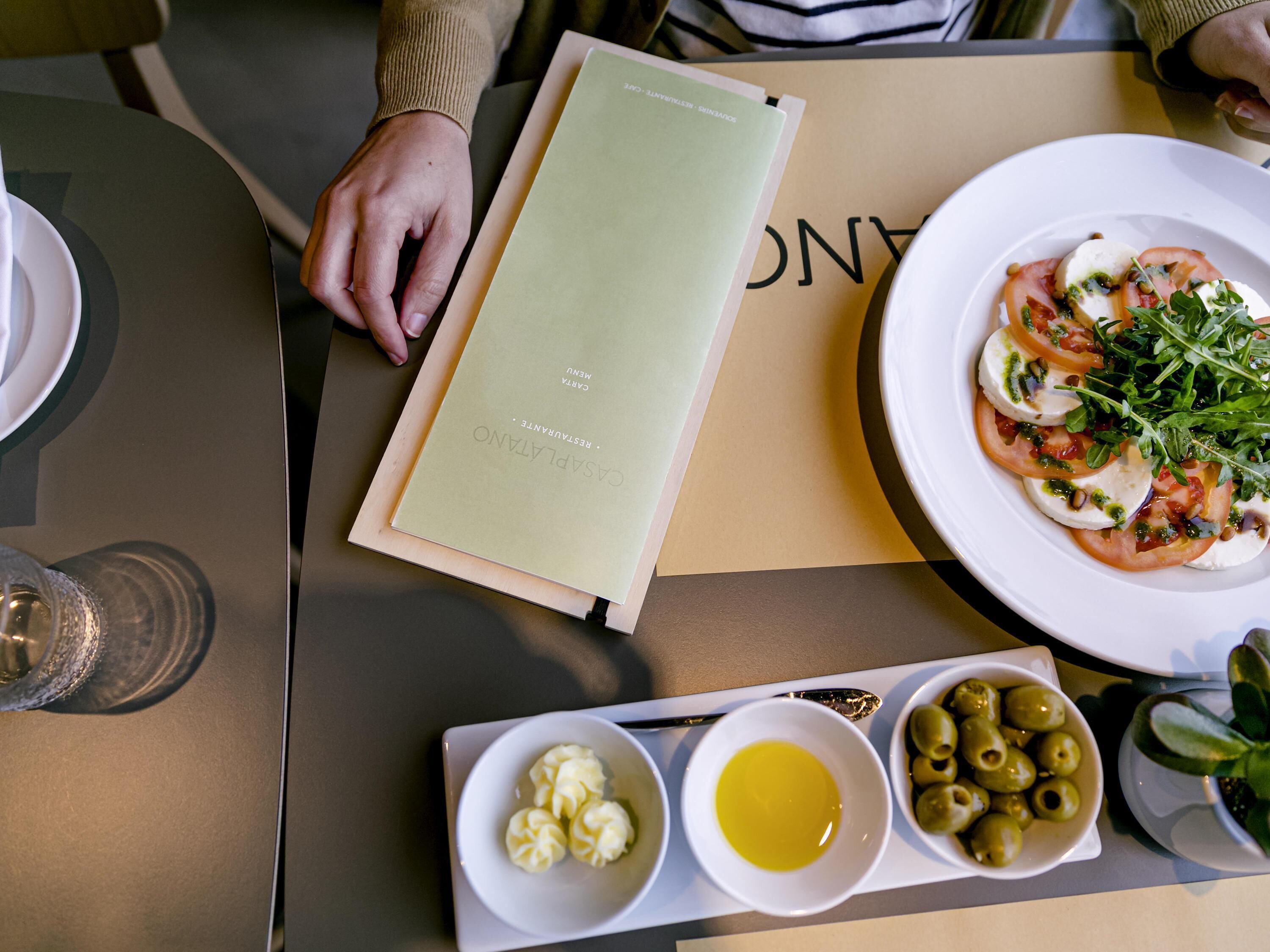 A menu, a plate with food, and a tray with olives and a bowl of sauce.