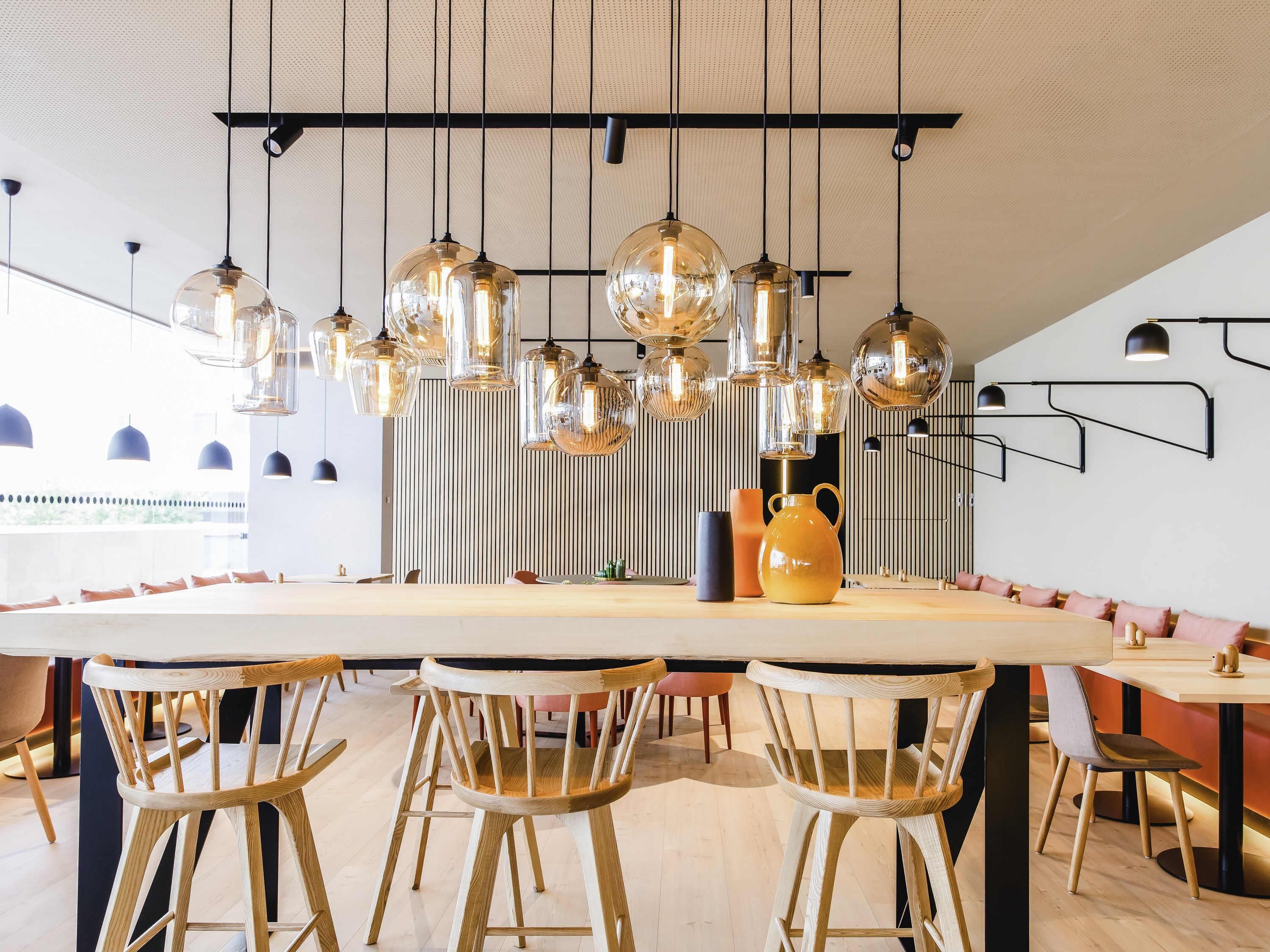 A modern dining area with a long communal table, wooden chairs, hanging pendant lights, and a decorative vase.