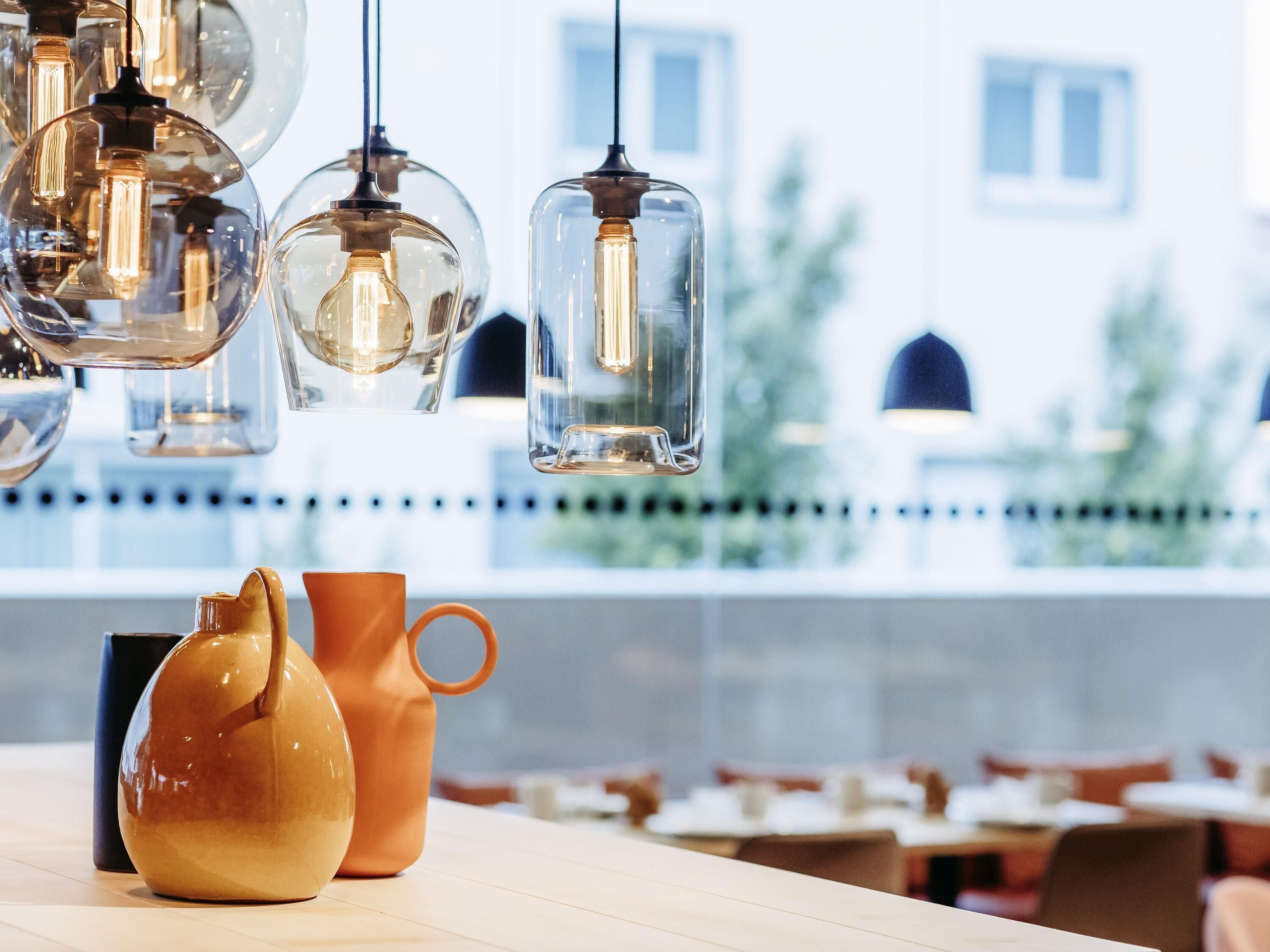 A restaurant interior with hanging pendant lights, a table with a vase and pitcher, and a view of buildings outside.