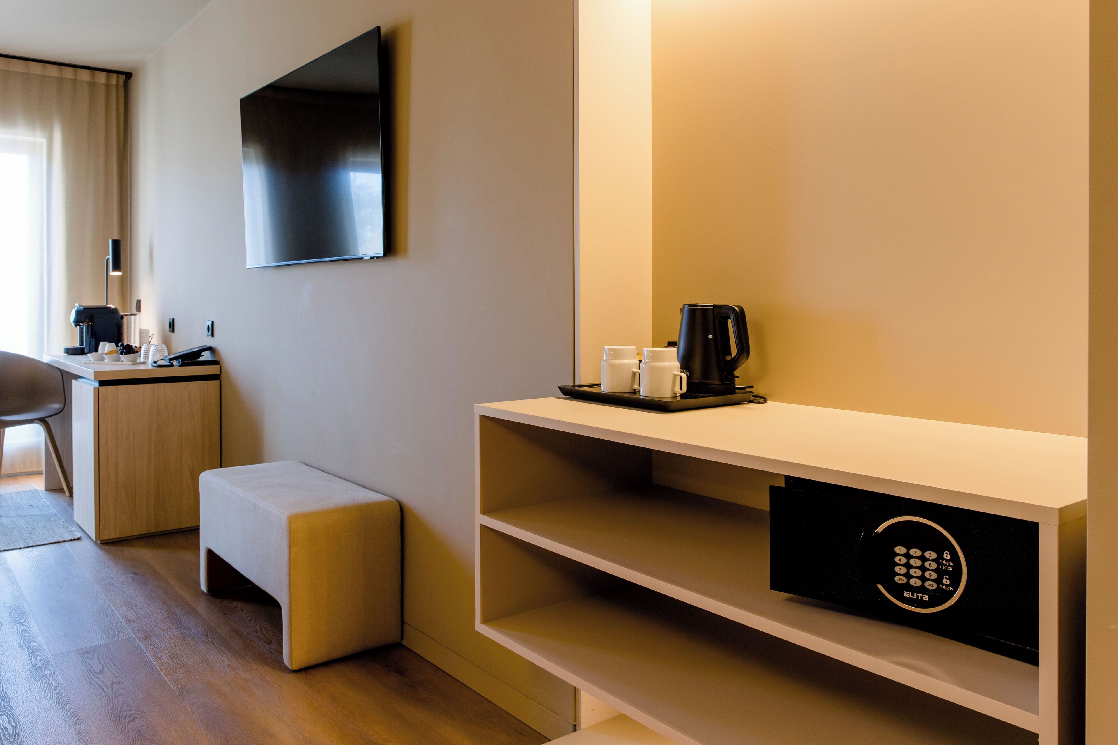 A hotel room with a flat-screen TV mounted on the wall, a coffee maker, and a tray with cups on a shelf.