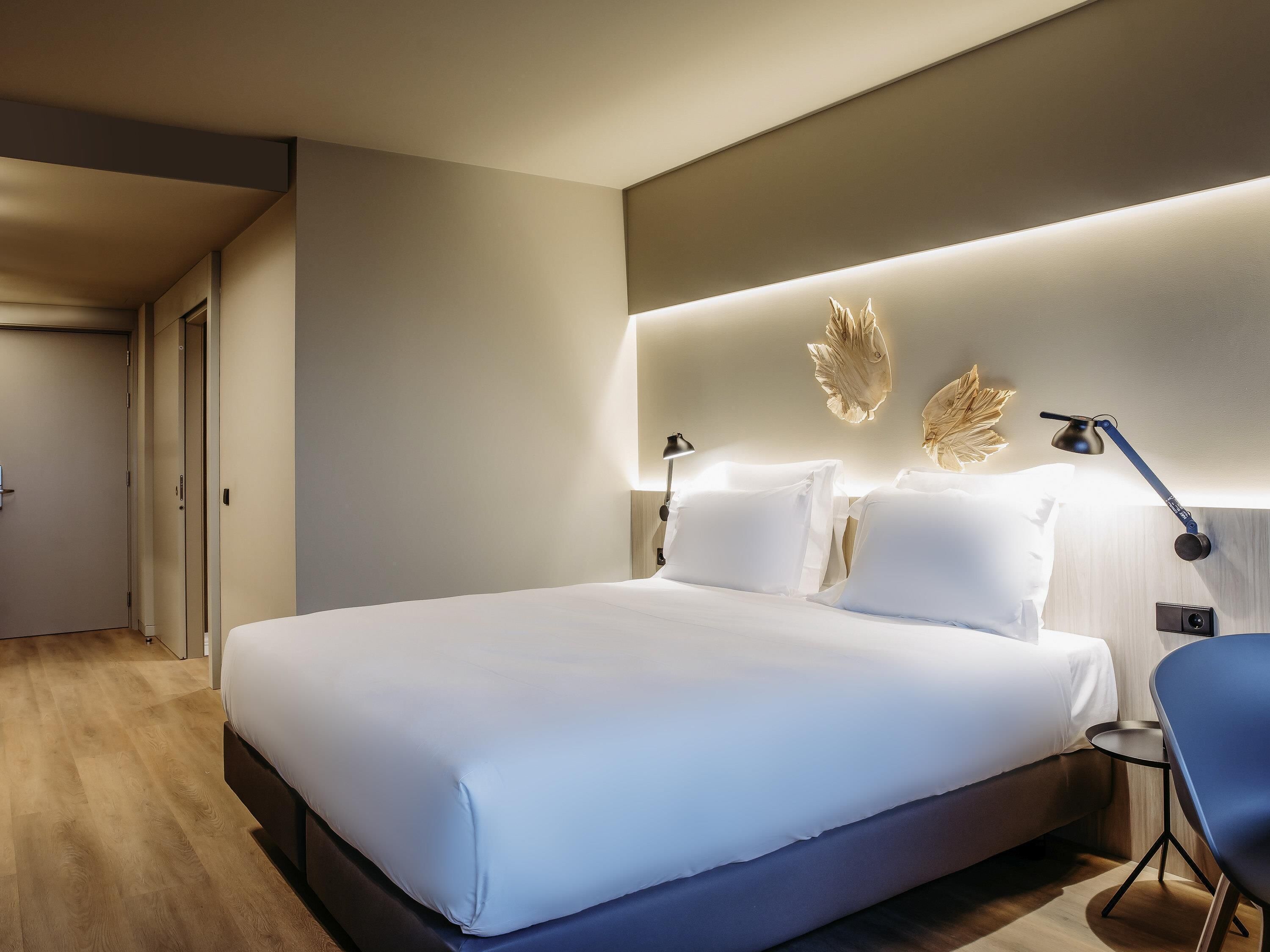 A modern hotel room with a large bed, bedside tables, a desk, and a wall-mounted lamp.