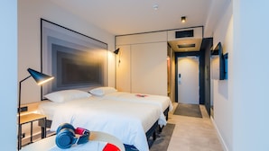 Una habitación de hotel moderna con dos camas, una mesita de noche con auriculares, una lámpara y una puerta que da a otra habitación.