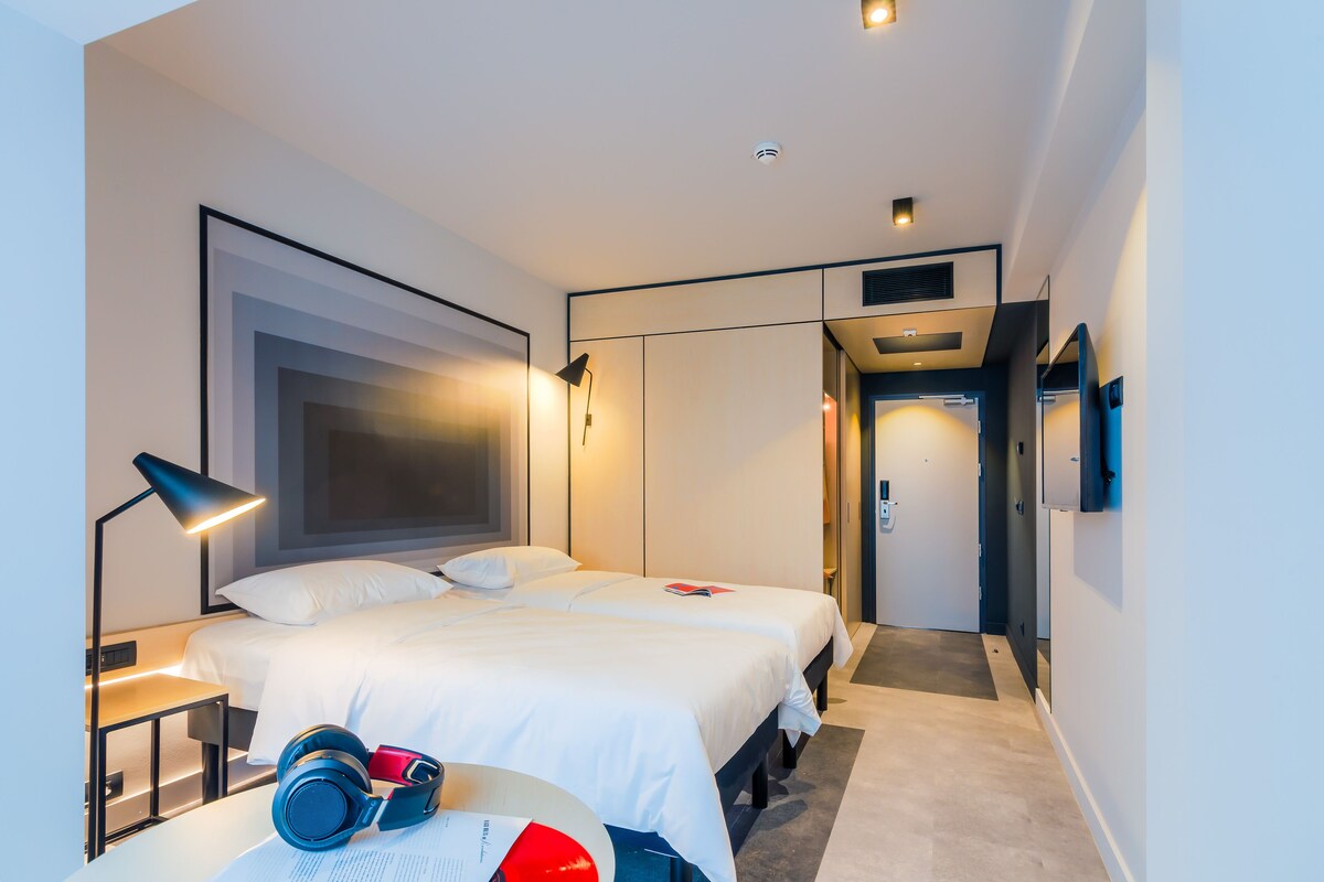 Una habitación de hotel moderna con dos camas, una mesita de noche con auriculares, una lámpara y una puerta que da a otra habitación.