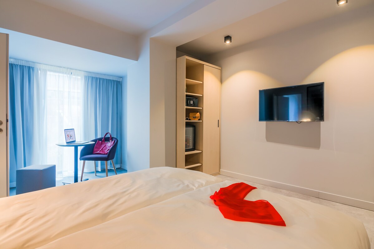 Habitación de hotel con cama, televisor montado en la pared, escritorio con silla y armario.