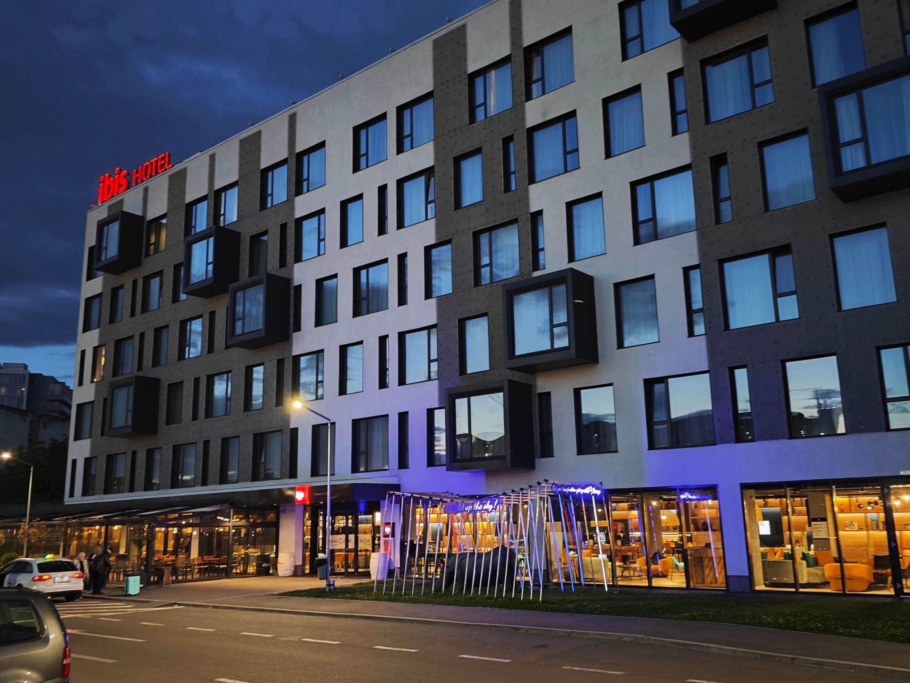 A modern hotel building with a prominent 'ibis HOTEL' sign, multiple windows, and a well-lit entrance area.