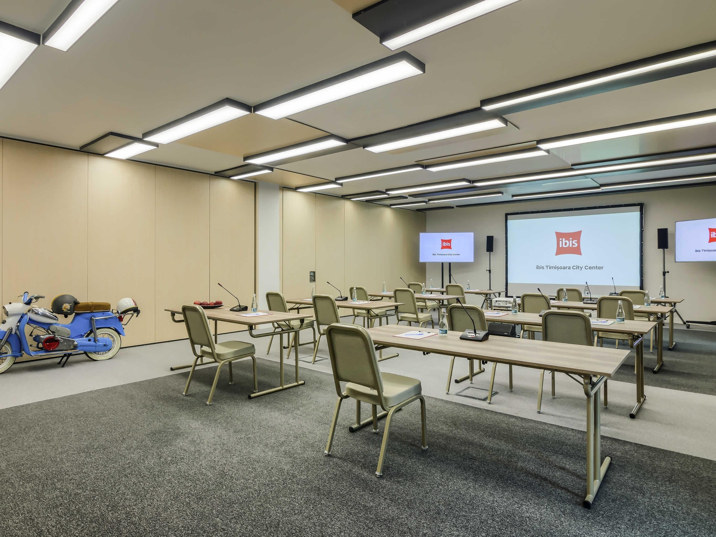 Un salón de conferencias con mesas, sillas, una pantalla de proyector y una motocicleta.