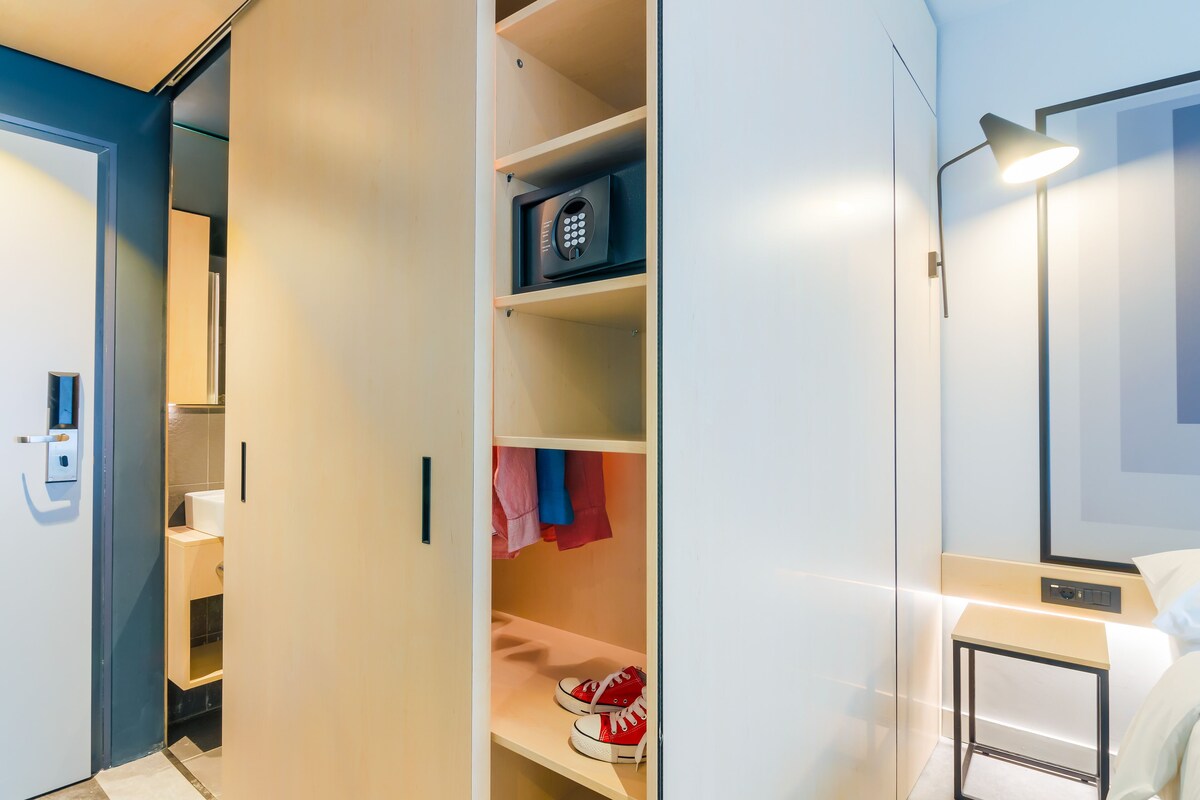 Una habitación de hotel moderna con vestidor, mesita de noche con lámpara y un espejo grande.