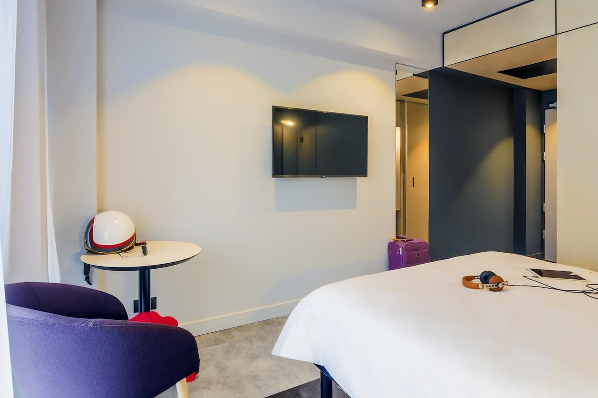 Habitación de hotel con cama, televisión montada en la pared, una mesita con un casco y una maleta, y una silla.