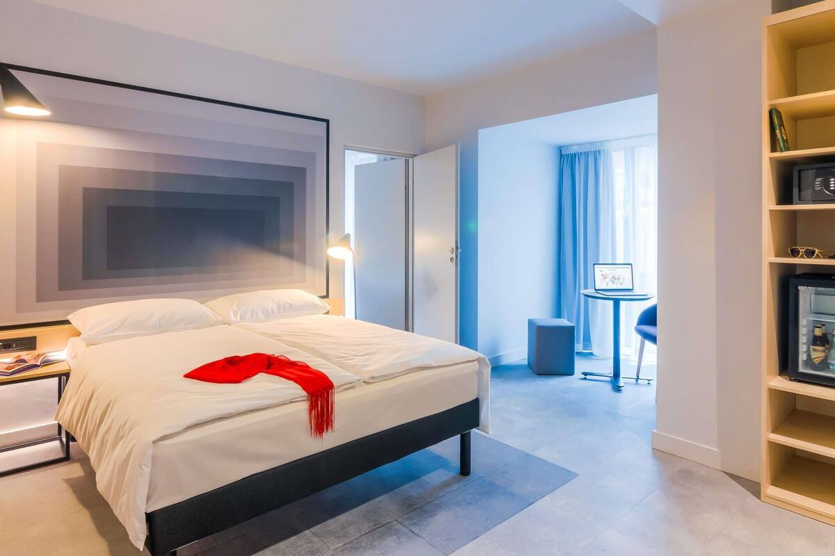Una habitación de hotel moderna con una cama grande, un escritorio, una estantería y vistas al exterior.