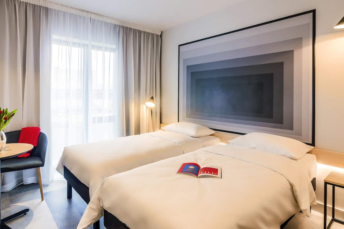 Habitación de hotel con dos camas, un escritorio, una silla, una obra de arte grande enmarco y una ventana con cortinas.