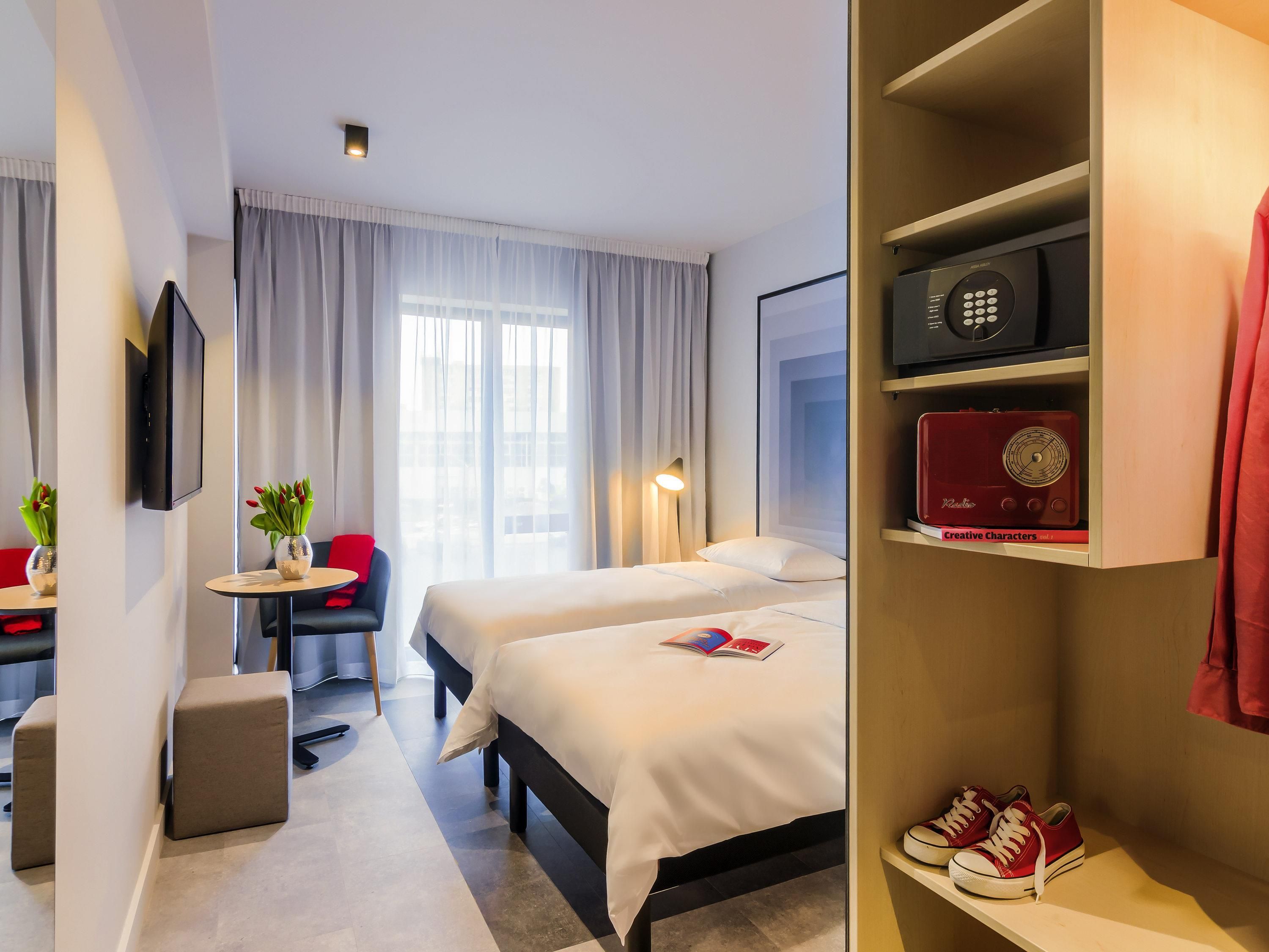 A hotel room with a bed, bedside table, a TV, a small table with a chair, a wardrobe, and a pair of shoes.