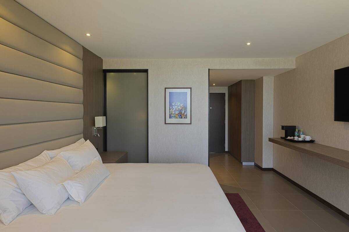 Ein Hotelzimmer mit einem großen Bett, einem Nachttisch mit Lampe, einem gerahmten Bild an der Wand und einem wandmontierten Flachbildfernseher.