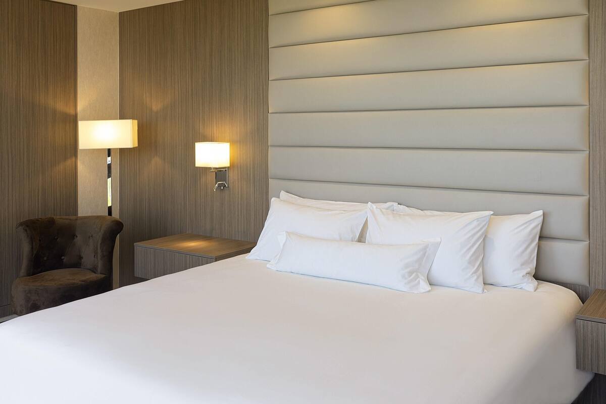Ein Hotelzimmer mit einem großen Bett, zwei Nachttischlampen, einem Sessel und einer Wandpaneele aus Holz.