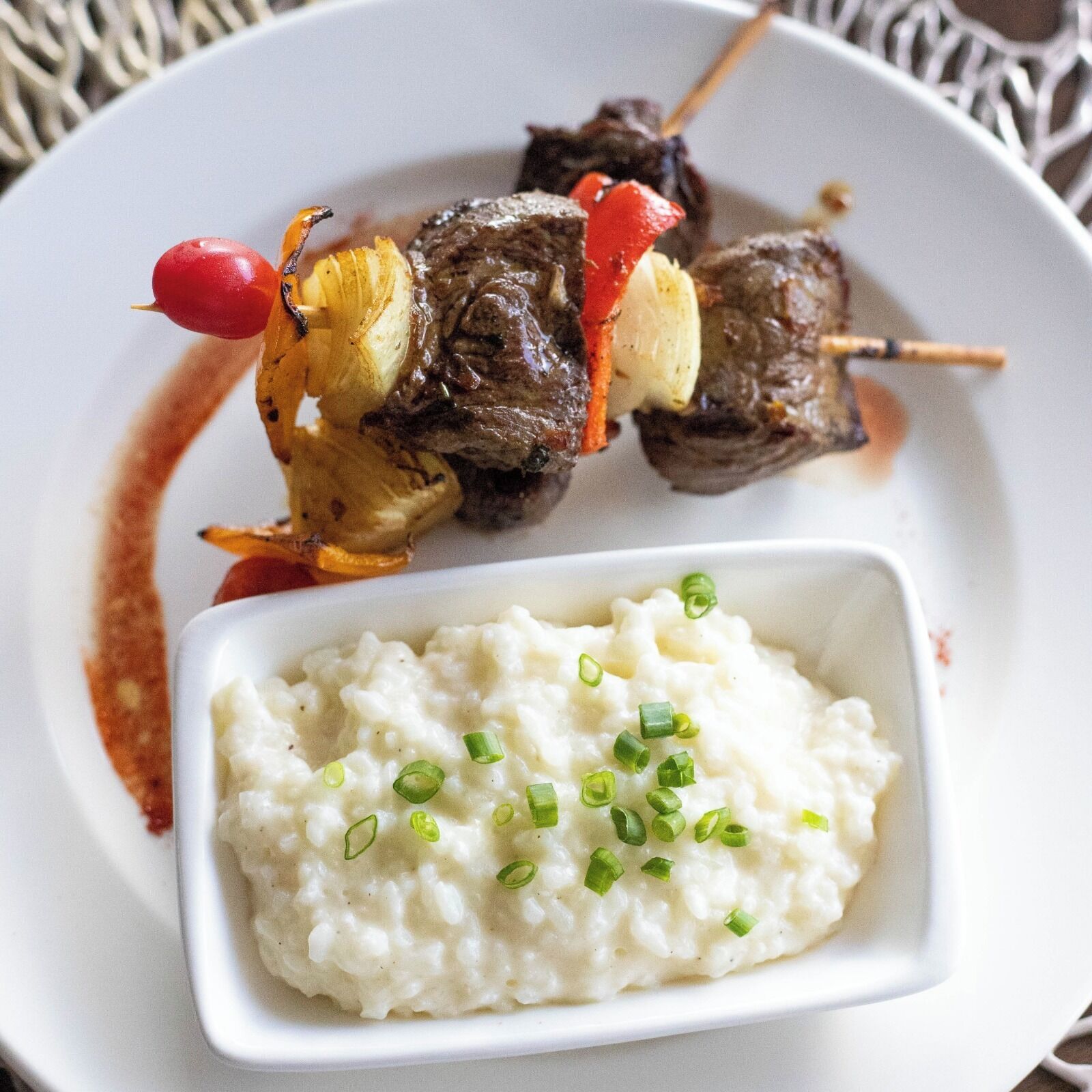 Un plato con brochetas de carne y verduras, acompañado de puré de papas cremoso adornado con cebollines.
