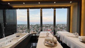 Habitación de hotel de gran altura con una bañera grande, dos camillas de masaje y vistas a la ciudad.