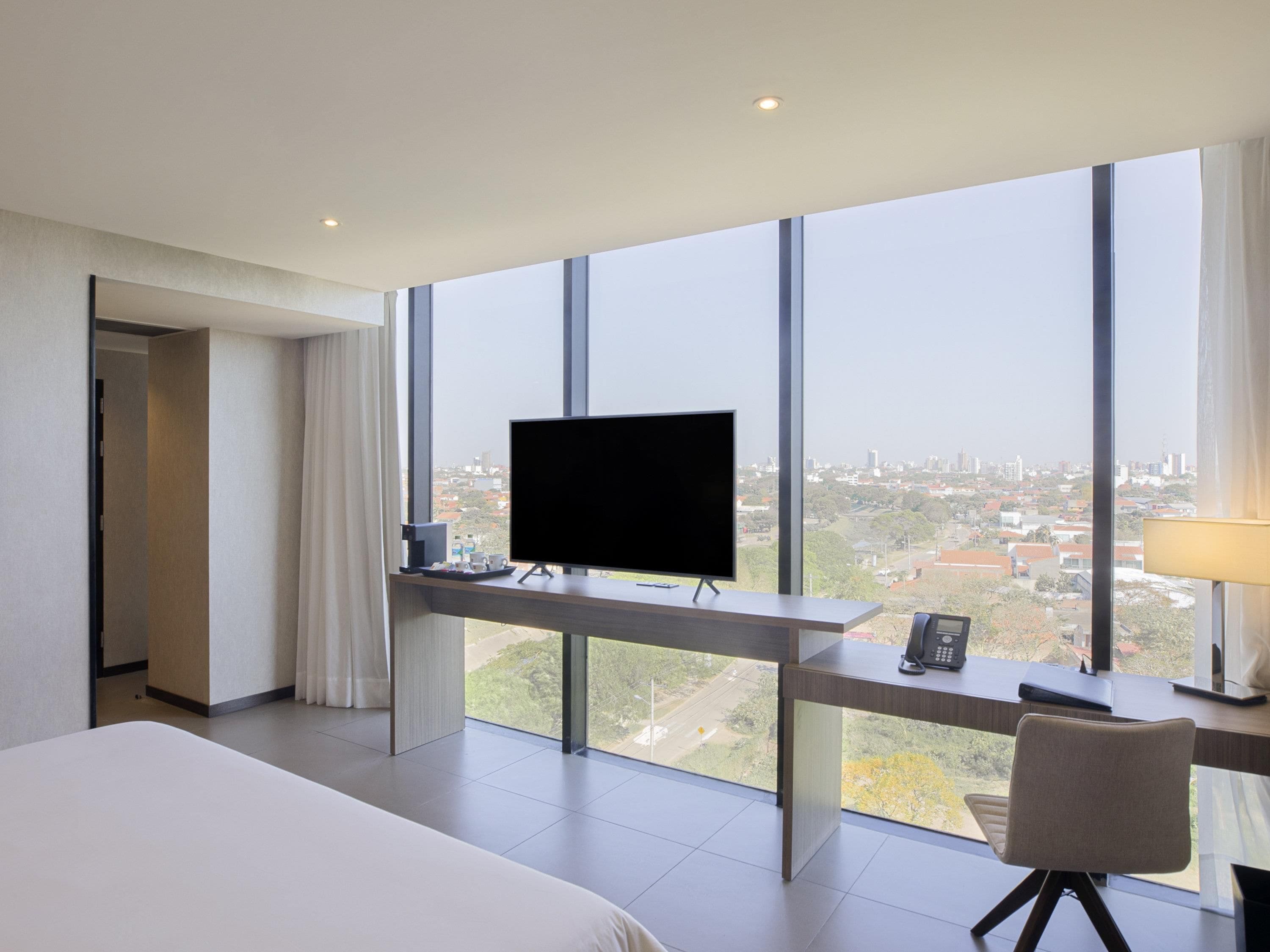 A hotel room with a large bed, a desk with a TV, and a view of a cityscape through large windows.