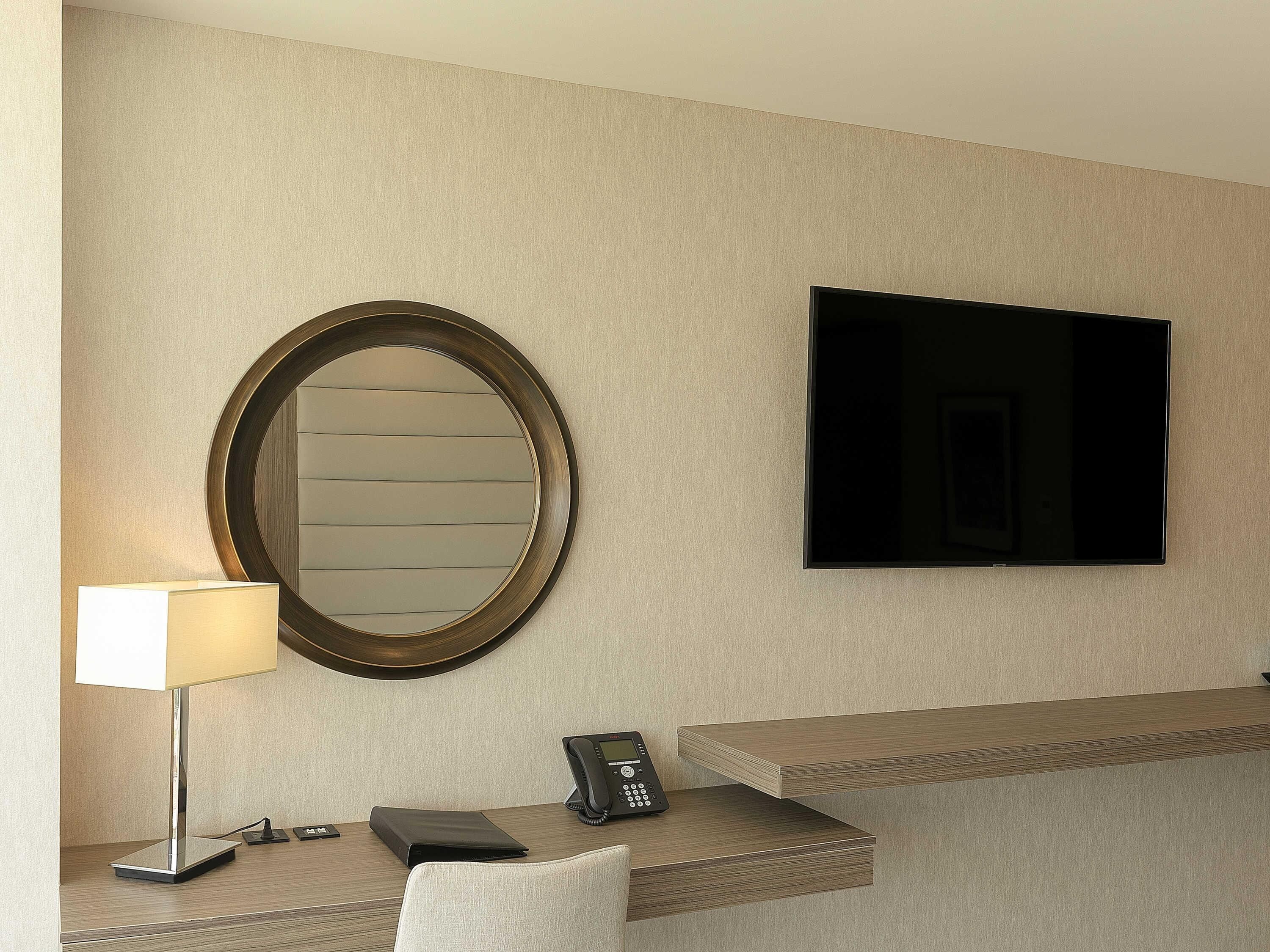 Une chambre d’hôtel dotée d’un miroir rond, d’un téléviseur à écran plat, d’un bureau avec une lampe, d’un téléphone et d’un banc rembourré.