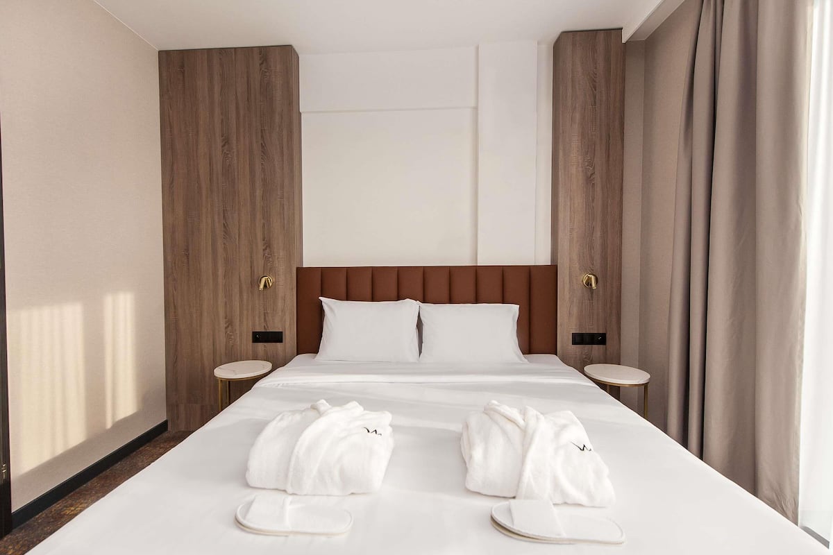 Une chambre d’hôtel avec un grand lit, deux tables de chevet, une tête de lit en bois et une vue sur l’extérieur.