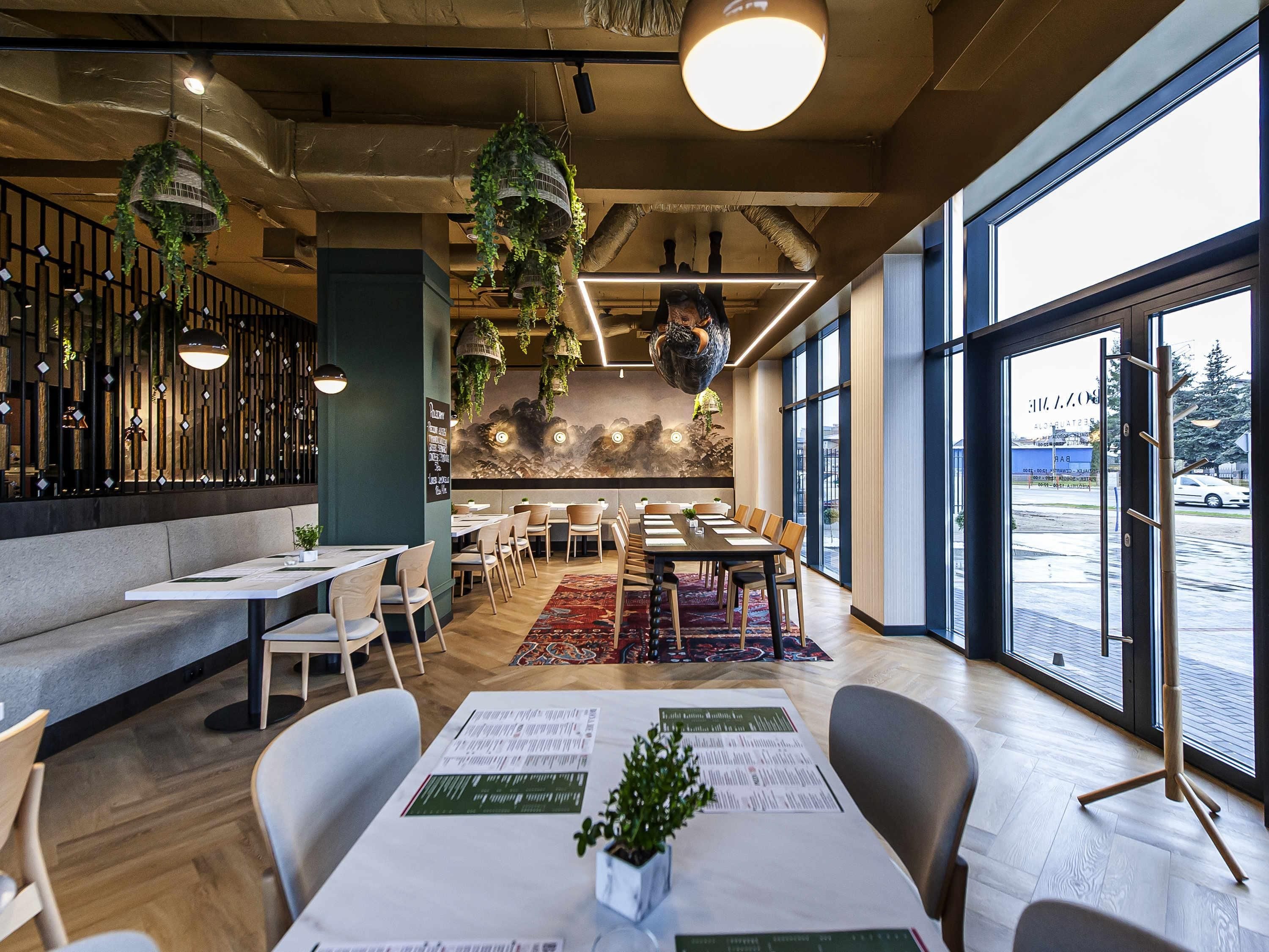 Restaurante com mesa comunitária grande, assentos individuais, plantas pendentes e janelas amplas.