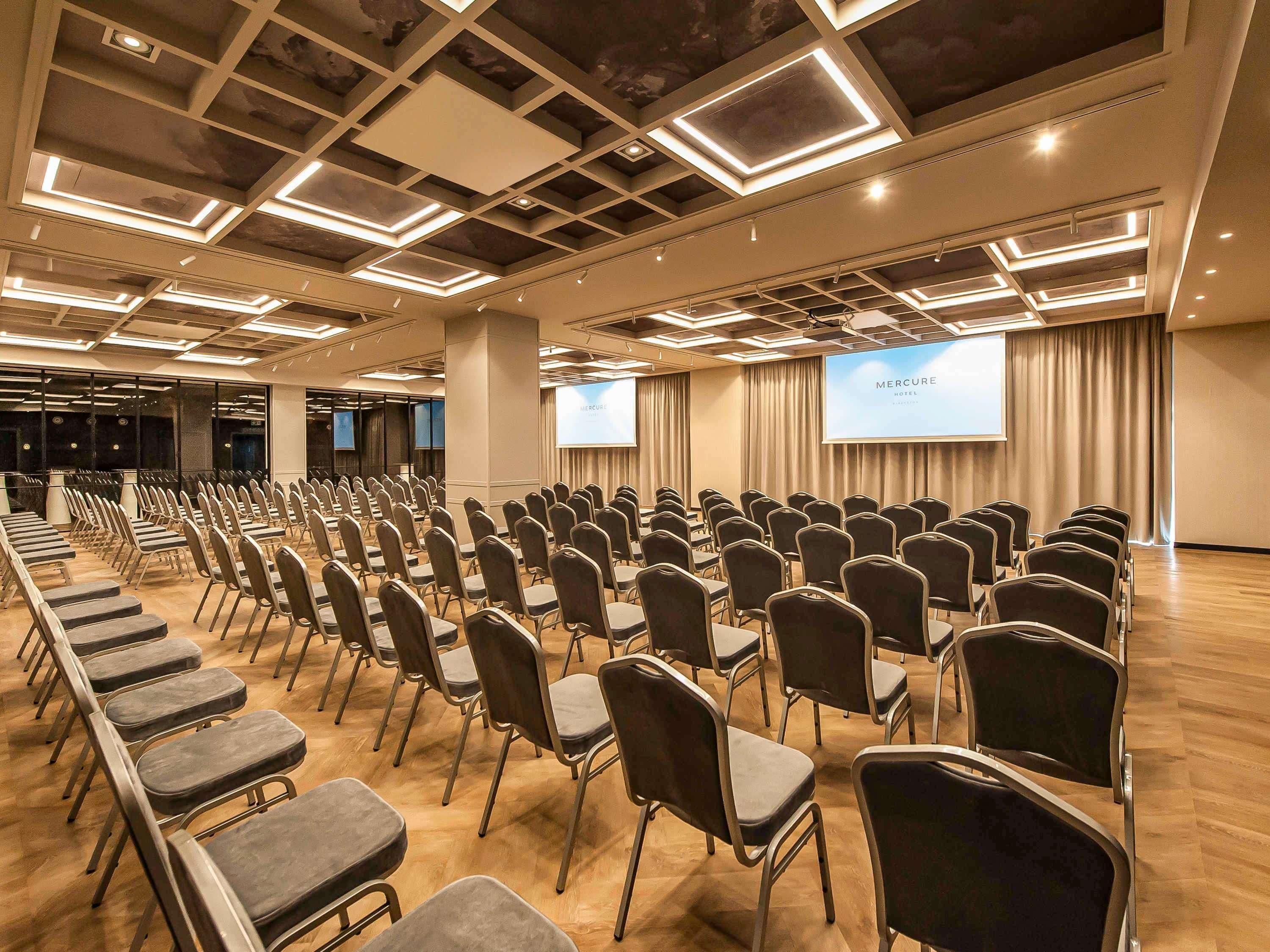 Ein Konferenzraum mit Reihen von Stühlen und einer Projektorleinwand. Ein großer Bildschirm zeigt 'Mercure Hotel'.