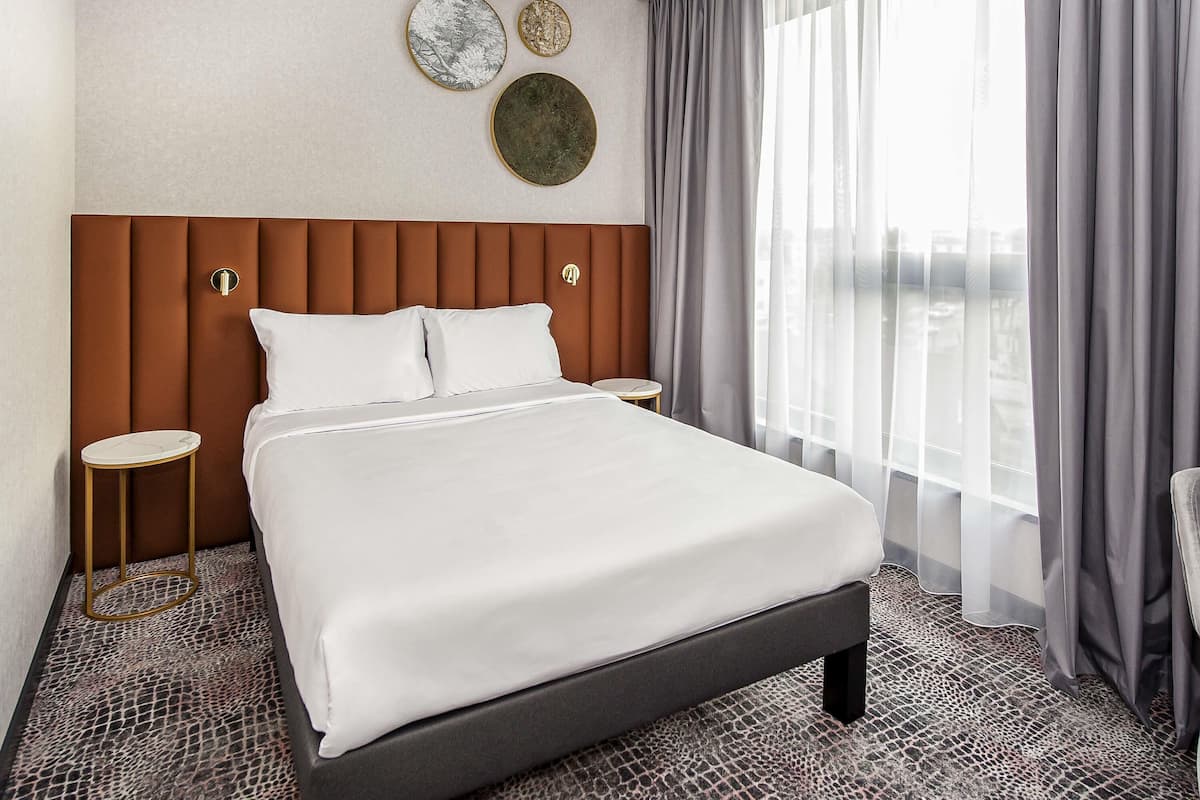 Une chambre d’hôtel avec un grand lit, deux tables de chevet, une tête de lit et une vue sur la ville à travers des rideaux transparents.