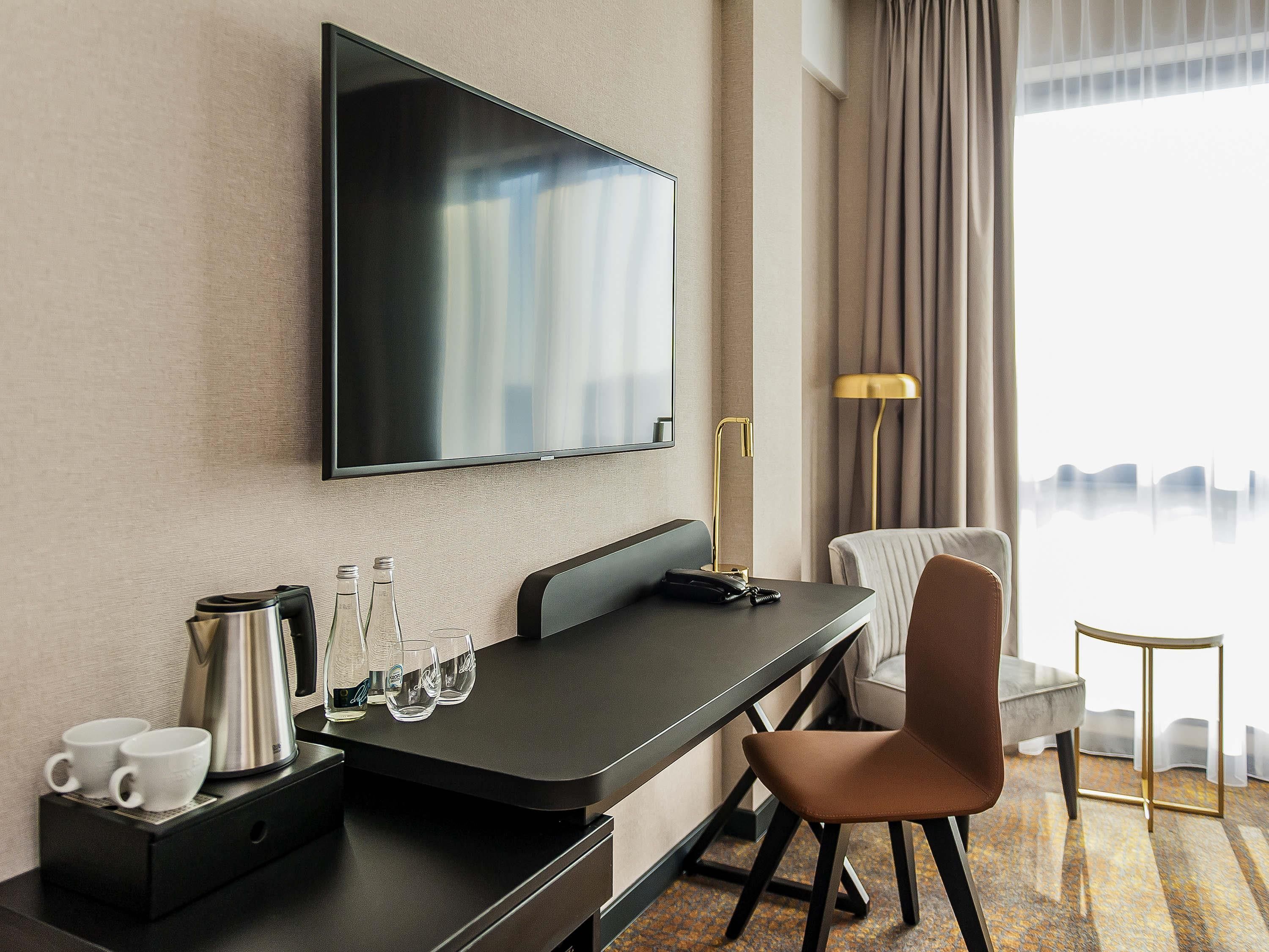 A modern hotel room with a flat-screen TV mounted on the wall, a desk with a coffee maker, glasses, and a telephone. There is a chair and a lamp beside the desk.