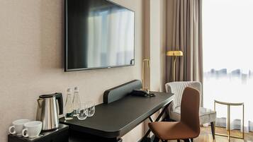 A modern hotel room with a flat-screen TV mounted on the wall, a desk with a coffee maker, glasses, and a telephone. There is a chair and a lamp beside the desk.