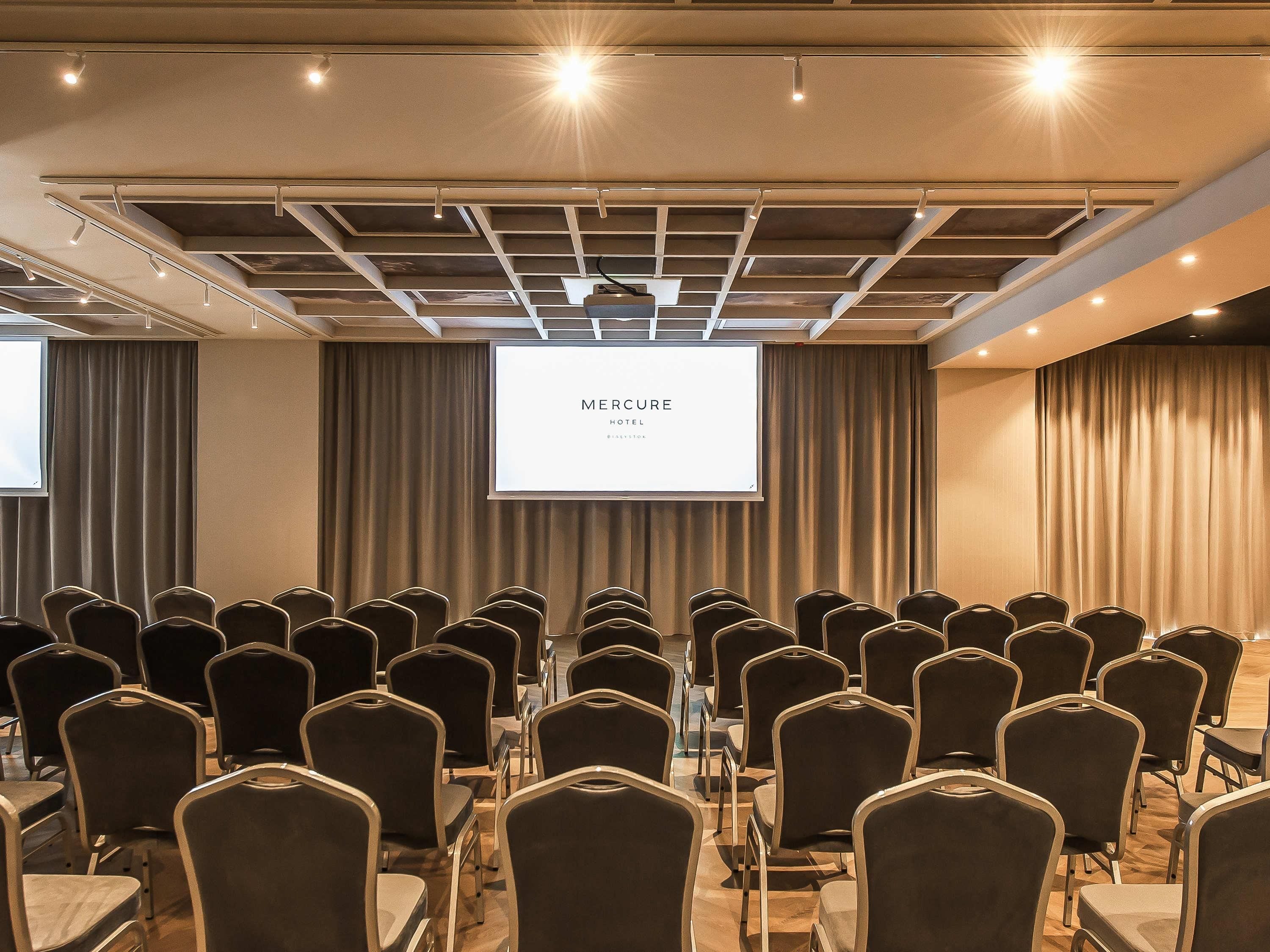 A conference room with rows of chairs, a projection screen, and a ceiling with recessed lighting.