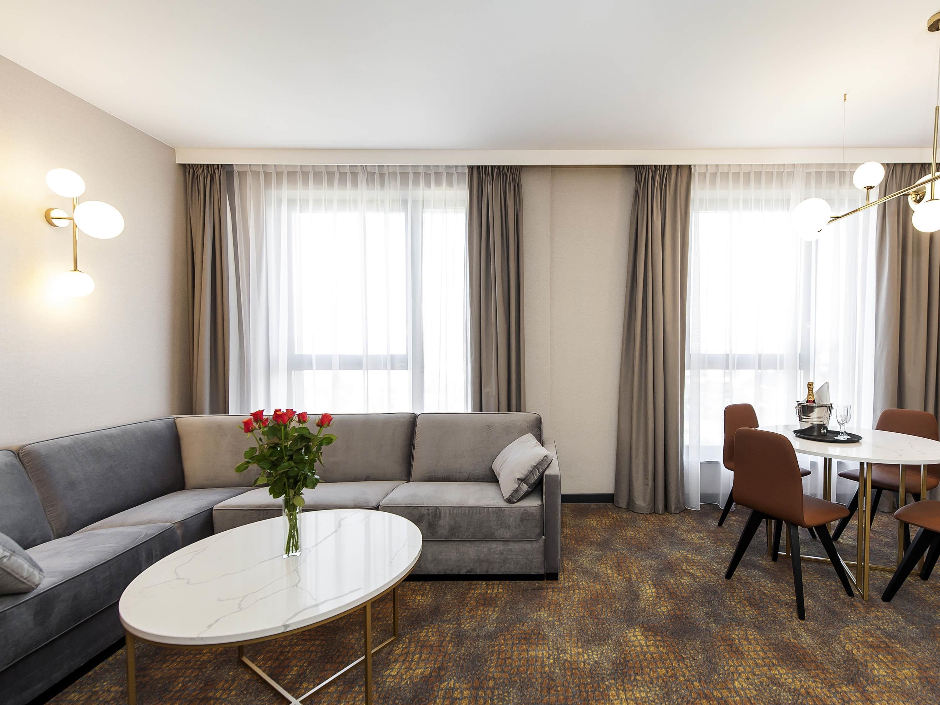 Et moderne hotelværelse med en grå modulsofa, et rundt hvidt sofabord, et spisebord med fire stole og en vase med røde roser.