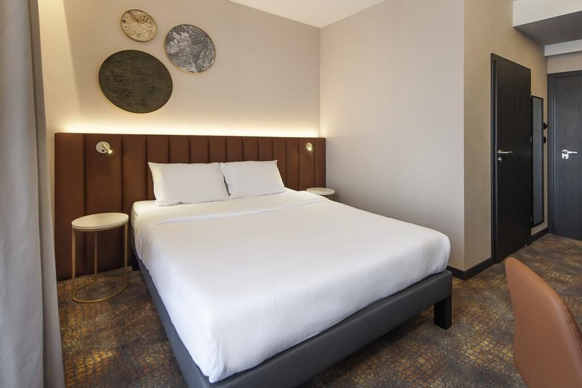 A hotel room with a bed, bedside tables, a door, and wall decor.