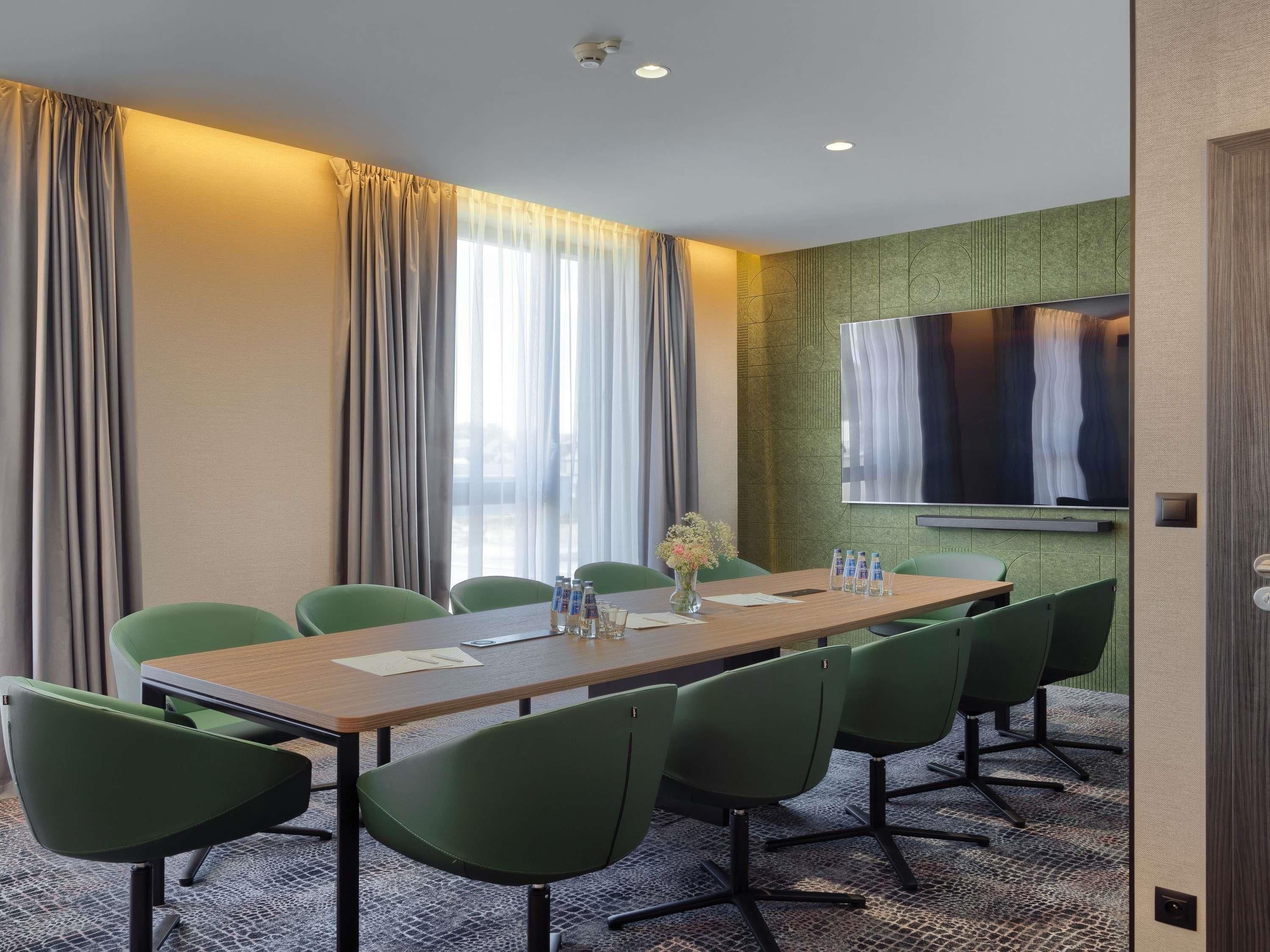 Un salón de conferencias con una larga mesa, seis sillas, un jarrón con flores y una pantalla instalada en la pared.