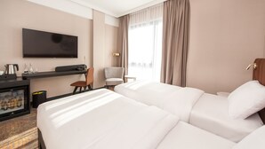 Habitación de hotel con una cama grande, un escritorio con televisor, minibar y vistas a la ciudad a través de amplios ventanales.