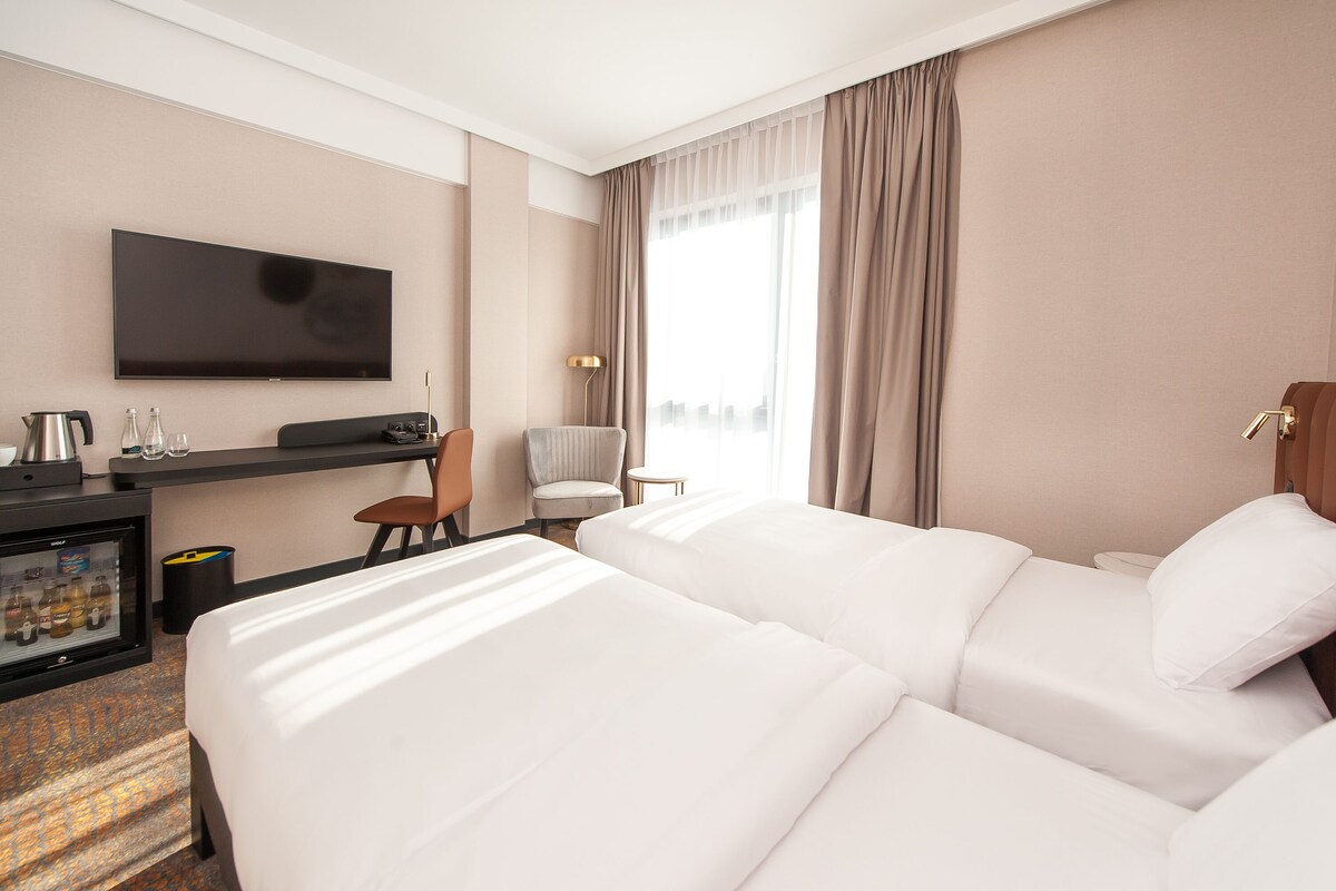 A hotel room with a large bed, a desk with a television, a minibar, and a view of the city through large windows.
